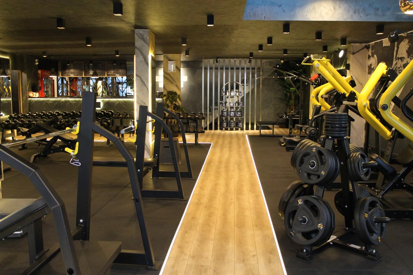 Space Gym: ışık şeritli ahşap yürüme yolu, iki yanında plaka ağırlıklar ve sarı çerçeveli makineler